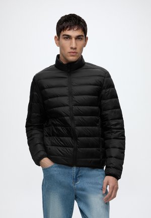 Jeune homme portant une veste noire matelassée avec fermeture éclair à l’avant et un jean bleu, debout avec une main dans la poche sur un fond uni.