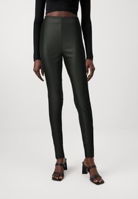 Leggings taille haute, vert foncé, avec une finition brillante. La coupe ajustée présente un matériau lisse et extensible avec des coutures minimales pour un look épuré.