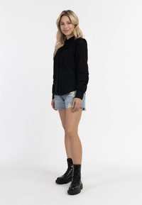 Zwarte lange mouwen shirt met textuur kant accenten, gecombineerd met versleten denim shorts en zwarte combat boots, staande tegen een witte achtergrond.