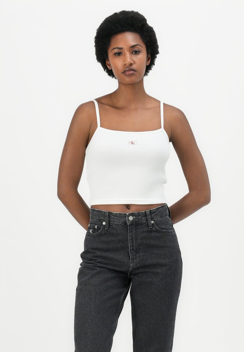 Weißes, geripptes Cropped-Top mit schmalen Trägern, das ein kleines, gesticktes rotes Logo zeigt. Kombiniert mit hochgeschnittenen, dunkelgrauen Jeans.