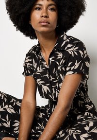 Schwarzes Kurzarmkleid mit beigem Blättermuster. Mit Kragen, Knopfverschluss und elastischem Bund. Strukturierter Stoff ist sichtbar.