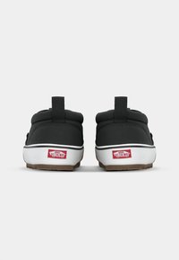 Sorte kanvas slip-on sko med polstret krage, hvit gummisåle og teksturert såle i tan. Har rødt "Vans Off the Wall"-logo på hælen.