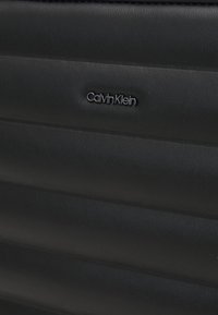 Černý kufr Calvin Klein se zvlněným designem a hladkým koženým povrchem. Logo je výrazně umístěno, což dodává jemný akcent.