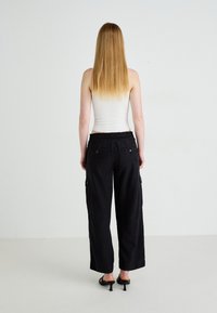 Zwarte cargo broek met een losse pasvorm, elastische tailleband en zijzakken. Geribbeld wit tanktop, lang steil haar. Effen achtergrond.