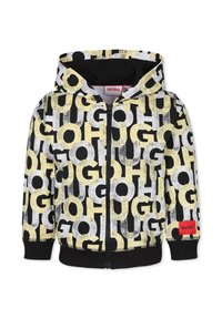 Sweat à capuche zippé noir, avec de grandes lettres graphiques jaunes et blanches. Étiquette rouge "HUGO" sur le devant. Texture douce, poignets côtelés.
