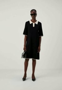 Μαύρο φόρεμα με κοντά μανίκια, λευκό γιακά και ευθεία σιλουέτα. Φορεμένο με μαύρες μυτερές γόβες και μια μικρή τσάντα, με κομψές υφές.