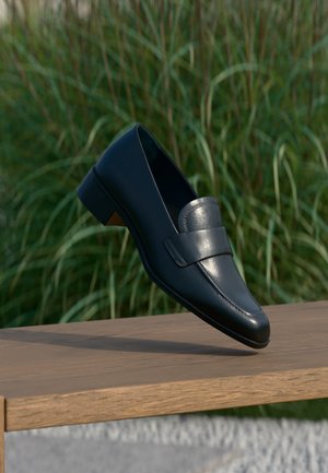 Mocassin en cuir noir à talon bas, en équilibre sur le bord d'un banc en bois à l'extérieur, avec de l'herbe verte et haute en arrière-plan.