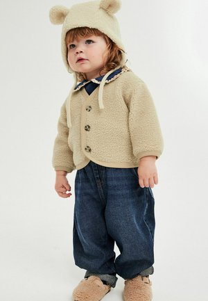 Beige cardigan i fleece med rund hals og knaplukning, kombineret med mørke denimbukser. Matchende beige hat med bjørneører og bløde hjemmesko.