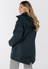 Donkergroene geïsoleerde jas met capuchon, met een gladde textuur en ritssluitingen in de zakken. De zoom heeft een lichte split voor meer bewegingsvrijheid.