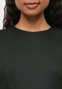 Detailní záběr dolní části obličeje, krku a horní části hrudníku osoby, která má na sobě jednoduchý černý top a vlnité tmavé vlasy viditelné po stranách.