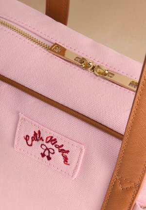 Różowa torba na ramię z płótna z złotym zamkiem błyskawicznym, brązowymi akcentami i czerwoną wyszywaną metką z nazwą marki.