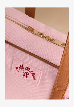 Różowa torba na ramię z płótna z złotym zamkiem błyskawicznym, brązowymi akcentami i czerwoną wyszywaną metką z nazwą marki.
