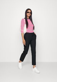 Red and white striped long-sleeve top, black tailored pants, white sneakers, and a textured white bag. Model poses against a light background.