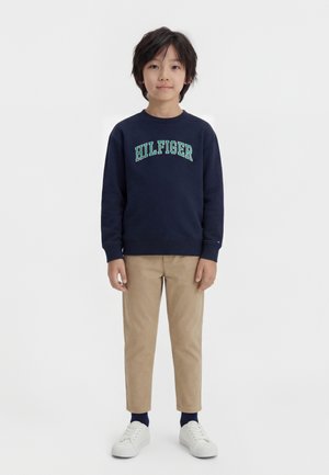 Child with black hair stands facing forward, wearing navy sweatshirt with "HILFIGER" text, beige pants, dark socks, and white sneakers against a white background.