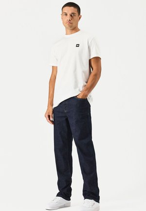 Junger Mann steht mit einer Hand in der Tasche seiner marineblauen Jeans, trägt ein weißes T-Shirt und weiße Sneakers vor einem einfarbigen Hintergrund.