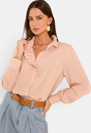 Vrouw met lang donker haar, die een roze geruite blouse met ruches draagt, blauwe hoge taille broek, bruine riem en gouden oorbellen, poserend met hand op heup.