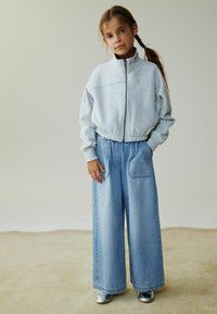 Svijetlosiva majica na patentni zatvarač s visokim ovratnikom, uparena s širokim plavim trapericama i sjajnim srebrnim cipelama.