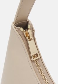 Beżowa skórzana torba o gładkiej fakturze, wyposażona w złoty zamek błyskawiczny oraz ozdobny suwak z logo, prezentująca minimalistyczny design.