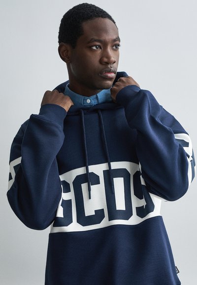 Νεαρός άντρας ρυθμίζει τον γιακά ενός ναυτικού μπλε φούτερ με μεγάλα λευκά γράμματα GCDS να διασχίζουν το στήθος και τα μανίκια.