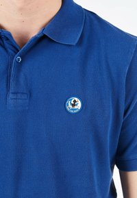 Polo blu realizzato in tessuto testurizzato con colletto. Presenta un logo circolare sul lato sinistro del petto, che raffigura un'anatra e un carrello.