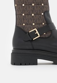Svart läderankelboot med en texturerad brun tyg med geometriskt mönster, gyllene fyrkantiga nitar och detalj med spänne på sidan.
