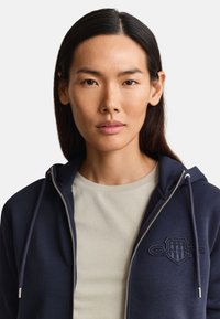 Nuori nainen, jolla on pitkät tummat hiukset, pukeutuu beigeen paitaan ja tummansiniseen vetoketjulliseen huppariin, jossa on kirjailtu logo, neutraali tausta.