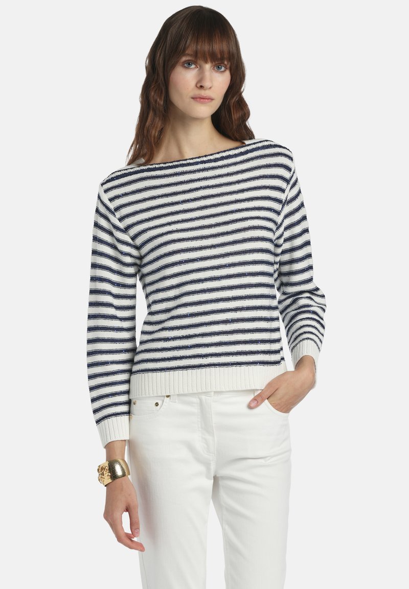 Femme portant un pull à manches longues rayé bleu marine et blanc, un pantalon blanc, et un large bracelet en or, avec une main dans la poche.