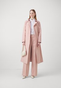 Lang rozenkleurige jas met een dubbelzijdig ontwerp, gecombineerd met een witte blouse en bijpassende rozenkleurige broek. Beschikt over een gestructureerde handtas en puntige schoenen.