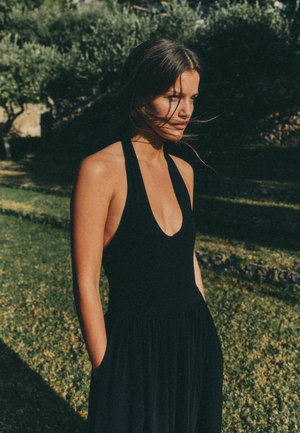 Naine musta varrukateta kleidis seisab vabalt väljas, käed taskutes, juuksed lehvimas ja taustal puud.