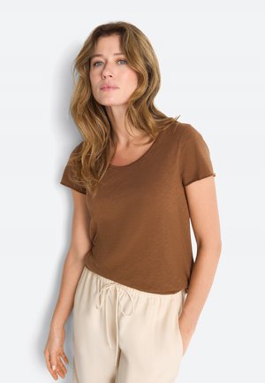 Kobieta o falujących jasnobrązowych włosach, ubrana w brązową koszulkę z krótkim rękawem i beżowe spodnie ze sznurkiem, stoi na tle białego tła.