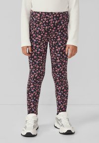 Kind trägt schwarze Leggings mit pinken, blauen und beigen Blumenmustern, ein weißes Langarmshirt und weiße klobige Sneaker und steht auf einem grauen Boden.
