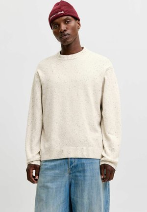 Homme portant un pull en maille crème tachetée, un jean bleu clair, un bonnet rouge en maille et une petite boucle d’oreille en anneau sur un fond gris uni.