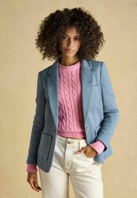 Tânără cu păr creț purtând un pulover roz tricotat cu model în relief, un sacou bleu deschis și pantaloni de culoare crem, pozând cu o mână în buzunar.