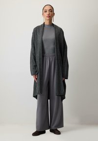 Dlhý, otvorený kardigán v tmavosivej pletenine, nosený cez šedý rebrovaný top a široké šedé nohavice. Hnedé látkové topánky dopĺňajú outfit.