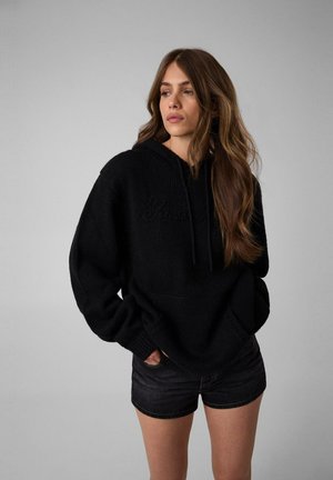 Giovane donna con lunghi capelli castani che indossa una felpa nera oversize e pantaloncini di jeans neri, in piedi davanti a uno sfondo grigio.