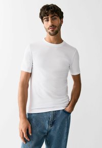 Vīrietis ar īsiem cirtainiem matiem un bārdu, tērpies vienkāršā baltā t-kreklā un zilās džinsu biksēs, ar vienu roku kabatā, stāvot pret vienkrāsainu balto fonu.