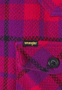 Nära bild av röd och lila rutigt ulltyg med en svart knapp och en liten svart etikett som står "Wrangler" i gult.