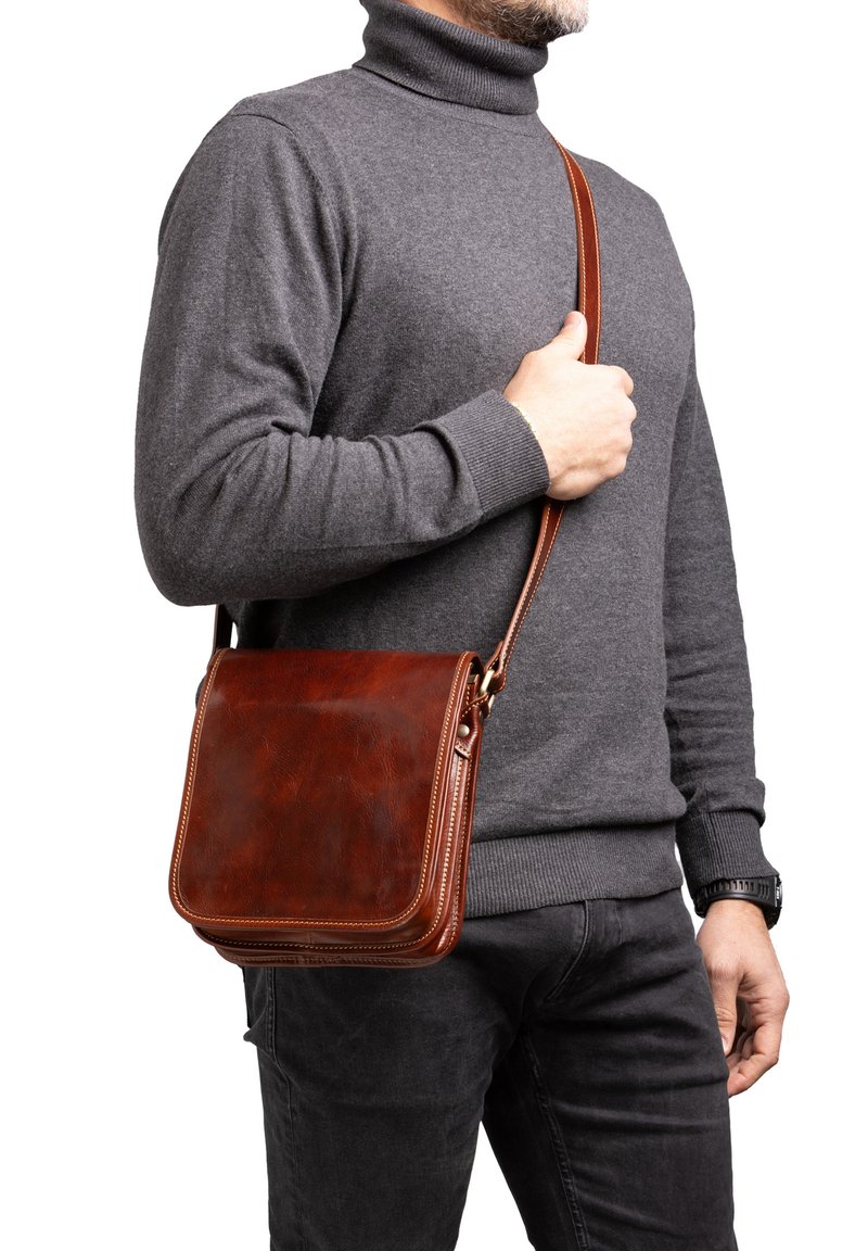 Borsa a tracolla in pelle marrone con forma rettangolare, texture liscia e una sottile tracolla regolabile, tenuta da una persona che indossa un dolcevita grigio.