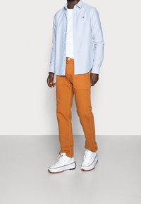 Lichtblauw overhemd over een wit T-shirt, gecombineerd met oranje broek en witte sneakers met zwarte zolen. Zichtbaar merkteken op het shirt.