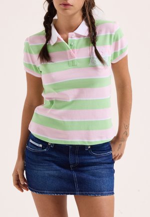 Donna che indossa una polo a righe rosa e verdi abbinata a una gonna di jeans blu scuro, in piedi con una mano in tasca.