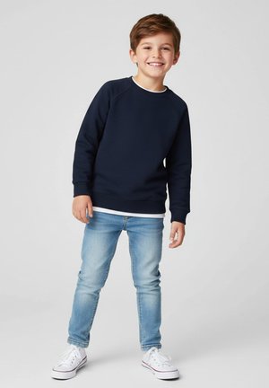 Chłopiec uśmiechający się, ubrany w granatową bluzę, jasnoniebieskie dżinsy i białe trampki, stojący na tle jednolitego jasnoszarego tła.