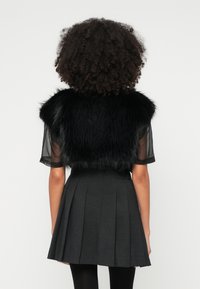 Colete preto de pelo sintético com mangas curtas transparentes, usado sobre uma saia preta justa e plissada. A textura é fofa e suave, adicionando dimensão ao conjunto.