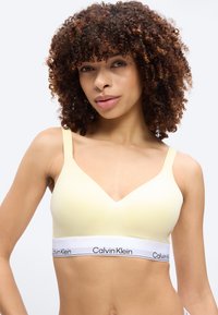 Γυναίκα με σγουρά μαλλιά, φορώντας ένα ανοιχτό κίτρινο bralette Calvin Klein με λευκή ελαστική ταινία με λογότυπο, ποζάροντας μπροστά σε απλό φόντο.