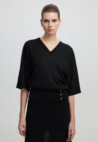 Pull noir à col v avec des manches chauve-souris, présentant un ourlet côtelé et trois accents de boutons dorés. Tissu tricot léger et design structuré.