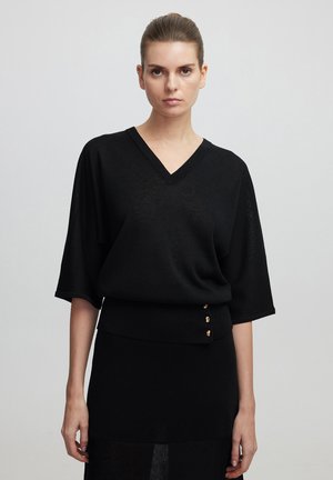Pull noir à col v avec des manches chauve-souris, présentant un ourlet côtelé et trois accents de boutons dorés. Tissu tricot léger et design structuré.