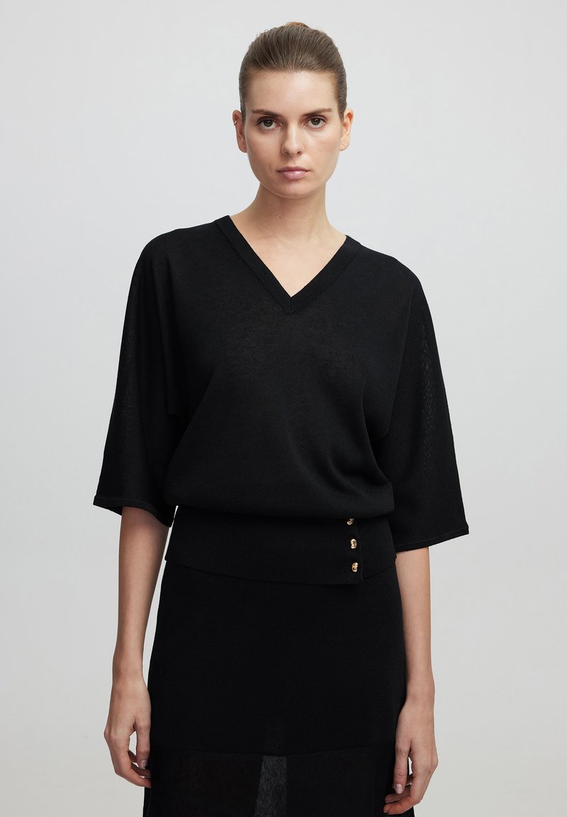 Pull noir à col v avec des manches chauve-souris, présentant un ourlet côtelé et trois accents de boutons dorés. Tissu tricot léger et design structuré.