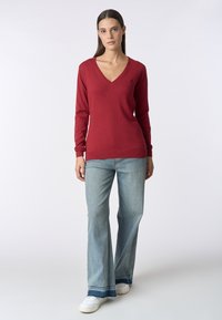Maglione rosso a maniche lunghe con scollo a V, orlo a costine e tessuto leggero, abbinato a jeans svasati azzurri e sneaker bianche.