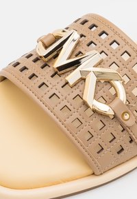 Sandale beige à bout ouvert avec une sangle carrée perforée et un détail de boucle métallique dorée sur une semelle crème.