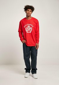 T-shirt rossa a maniche lunghe con logo "SOUTHPOLL" in bianco, abbinata a jeans scuri e scarpe da ginnastica bianche. La vestibilità è rilassata e casual.