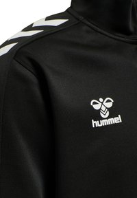 Zwarte sportjack met hoge kraag, gemaakt van een gladde stof. Bevat een wit logo en chevronaccenten op de schouders.